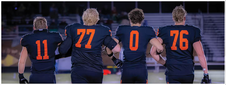 Four people wearing black sports uniforms with orange numbers are standing side by side on a field, holding hands or linking arms. Description generated by AI