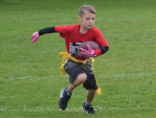 A child wearing a red shirt and pink gloves is running on grass while holding a football and wearing a yellow flag belt. Description generated by AI