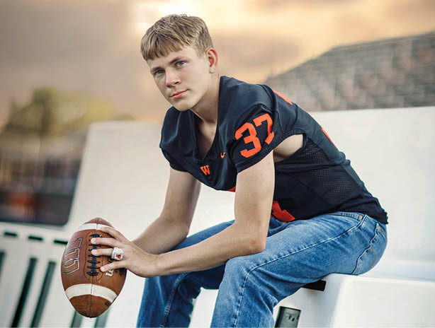 A person wearing a dark sports jersey with the number 37 is leaning forward outdoors with a blurred stadium and cloudy sky in the background. Description generated by AI