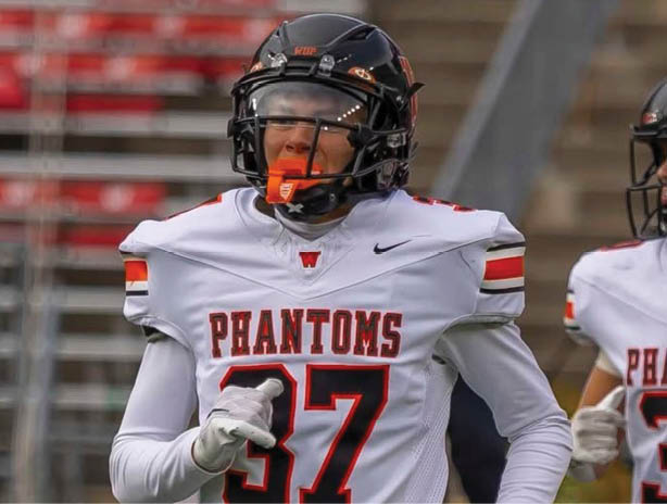 A person wearing a white football uniform with the word “PHANTOMS" and the number 37 is on a field with bleachers in the background. Description generated by AI