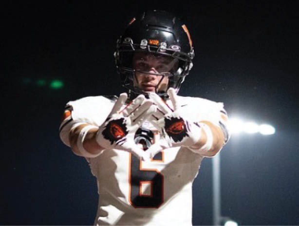 An adult wearing a white sports jersey and orange pants is jumping with arms raised on a football field at night while other players in similar uniforms stand in the background. Description generated by AI
