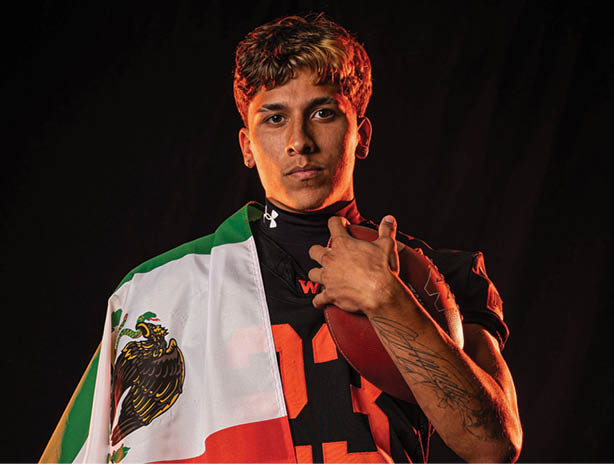 Person wearing a black sports jersey holding a football and draped with a Mexican flag over one shoulder against a dark background. Description generated by AI