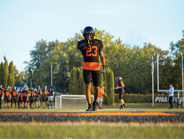 A person wearing a black and orange football uniform and helmet stands on a field with trees and a clear blue sky in the background. Description generated by AI