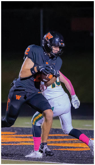 A person wearing a dark football uniform with the number 2 is holding a football tightly while being tackled by another person in a white and green uniform on a lit football field at night. Description generated by AI