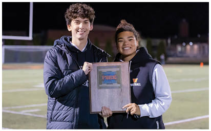 Two people smiling and holding a plaque on a sports field at night. Description generated by AI