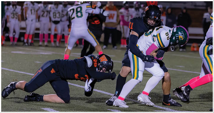 Two people in football uniforms are engaged in a tackle on a field while another player in a dark uniform is on the ground nearby. Description generated by AI