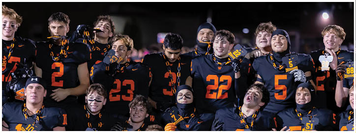 A group of people wearing black and orange football uniforms are gathered closely together, smiling and posing for a celebratory photo on a field at night. Description generated by AI