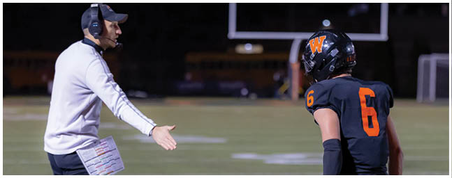 Two people stand on a football field, one wearing black athletic gear and the other holding a laminated play sheet. Description generated by AI