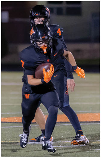 Two people in black football uniforms with orange gloves and helmets are on a field, with one holding a football and running while the other follows closely behind. Description generated by AI