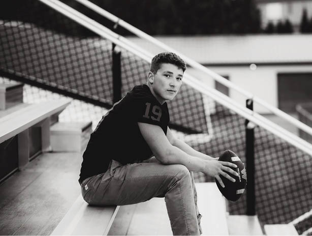 A person wearing a dark shirt and light pants is sitting on bleachers holding a football in an outdoor sports setting. Description generated by AI