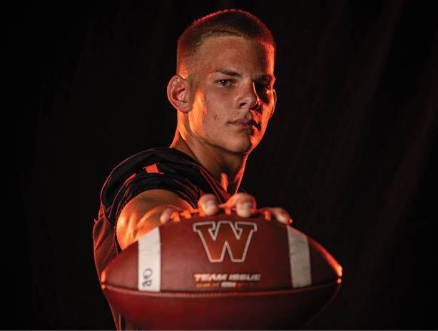 A person with short hair wearing an orange and black sports jacket with a large white “W" on the chest is looking directly at the camera against a dark background. Description generated by AI