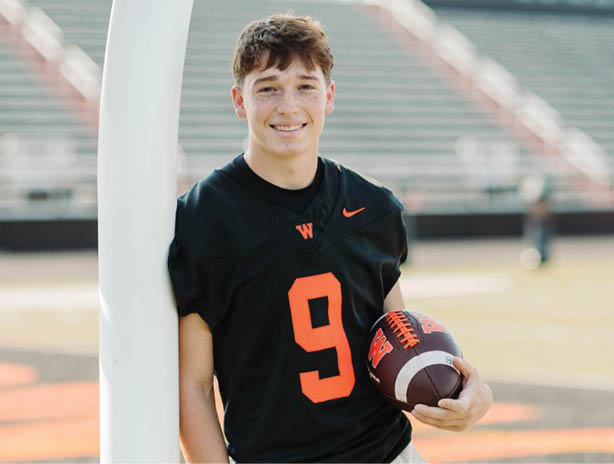 A person wearing a black sports jersey with the number nine is standing on a football field holding a football and leaning against a goalpost. Description generated by AI