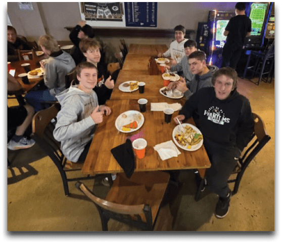 A group of people sitting around a long wooden table eating a meal together in a casual dining setting with a television and arcade machines in the background. Description generated by AI