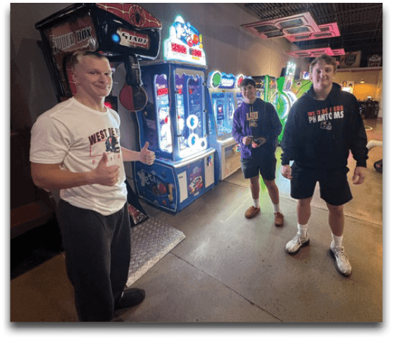 Three people stand indoors near arcade machines, with one person giving a thumbs-up and others casually dressed in shorts and sweatshirts. Description generated by AI