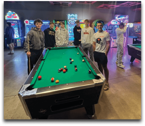 A group of people stand around a pool table in a dimly lit arcade room with neon lights and multiple gaming machines. Description generated by AI