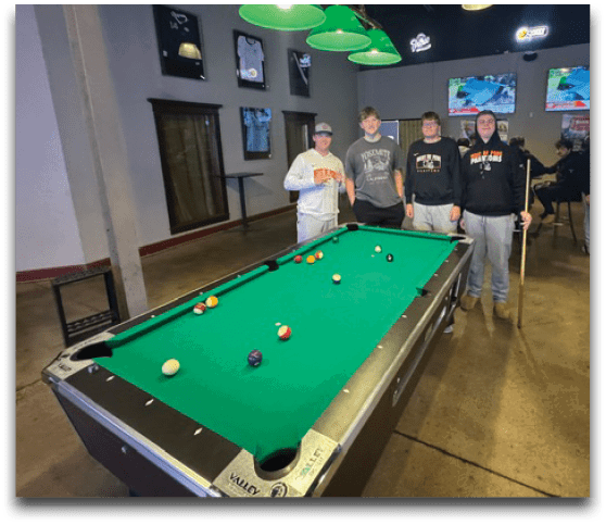 Four people stand around a pool table in a dimly lit room with green hanging lights and two televisions on the wall showing a sports game. Description generated by AI