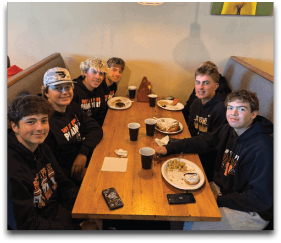 Four people with casual clothing and varied hairstyles are sitting closely together at a wooden table in a cozy booth under a hanging industrial-style light, with plates of food and drinks in front of them and framed photos on the wall behind. Description generated by AI