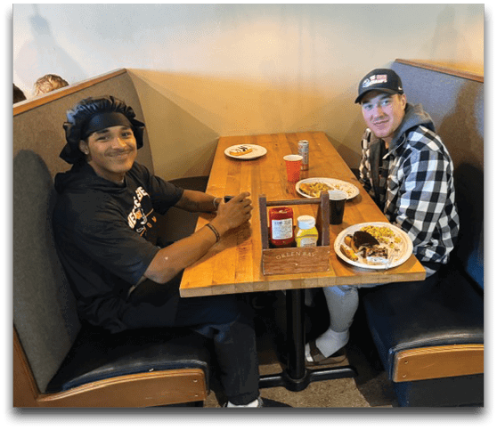 Two people sit at a wooden booth table in a casual dining setting with plates of food and condiments on the table. Description generated by AI