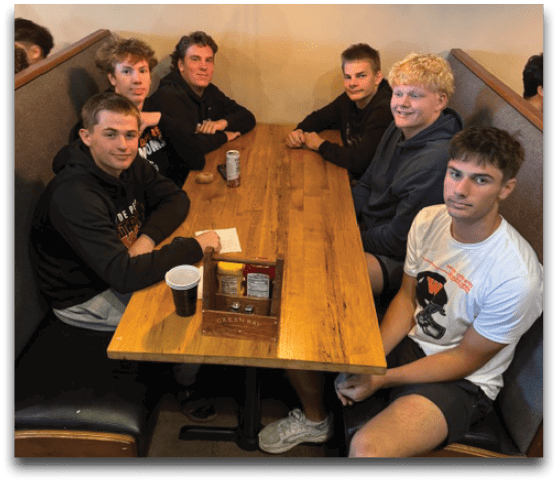 Seven people wearing casual clothing are sitting around a wooden table in a booth under a hanging light in a relaxed indoor setting. Description generated by AI