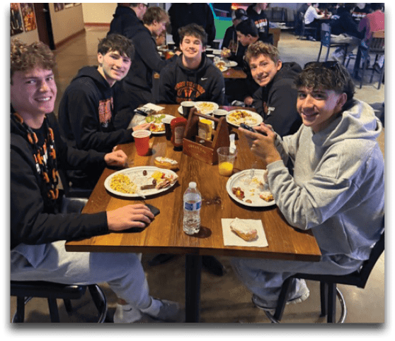 Five people wearing casual hoodies are sitting around a wooden table in a restaurant, smiling and eating food from plates with drinks and condiments on the table. Description generated by AI