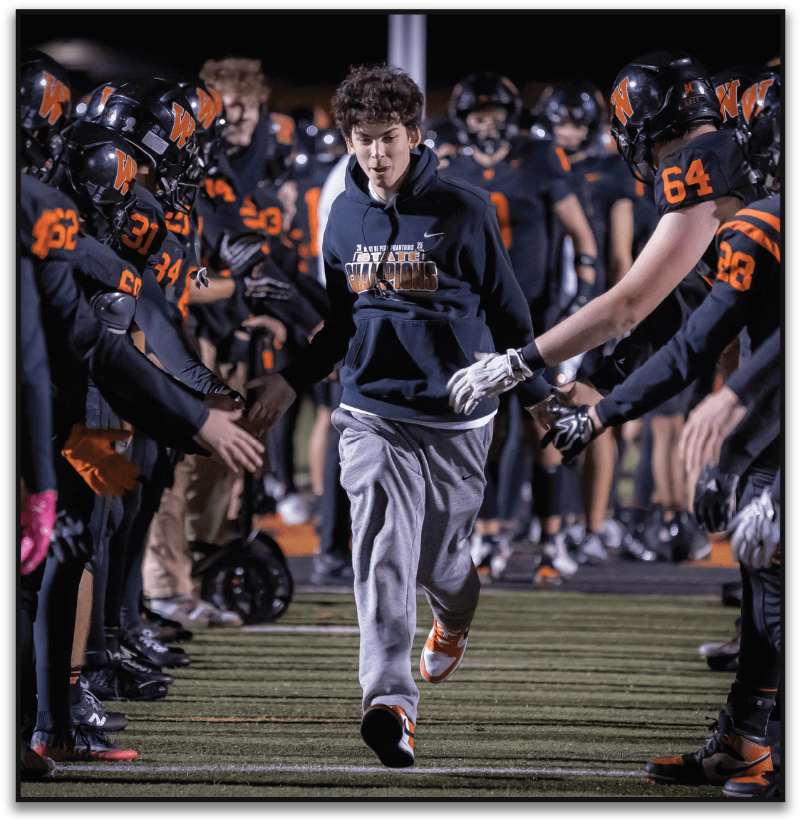 A person wearing a dark hoodie and gray sweatpants runs between two lines of people in black and orange football uniforms reaching out their hands on a sports field at night. Description generated by AI