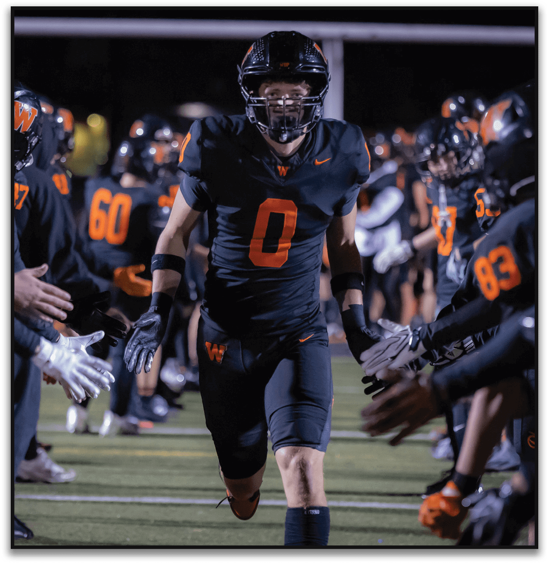 An athlete wearing a black football uniform with the number zero runs through a line of teammates giving high fives on a lit football field at night. Description generated by AI