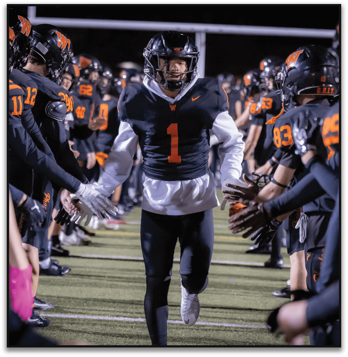 A person wearing a black football uniform with the number one runs down a field between two lines of teammates in matching uniforms reaching out to high-five. Description generated by AI
