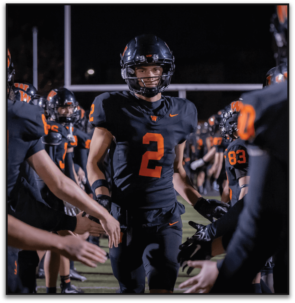 A person wearing a black football uniform with the number 2 is walking through a line of teammates giving high fives on a football field at night. Description generated by AI