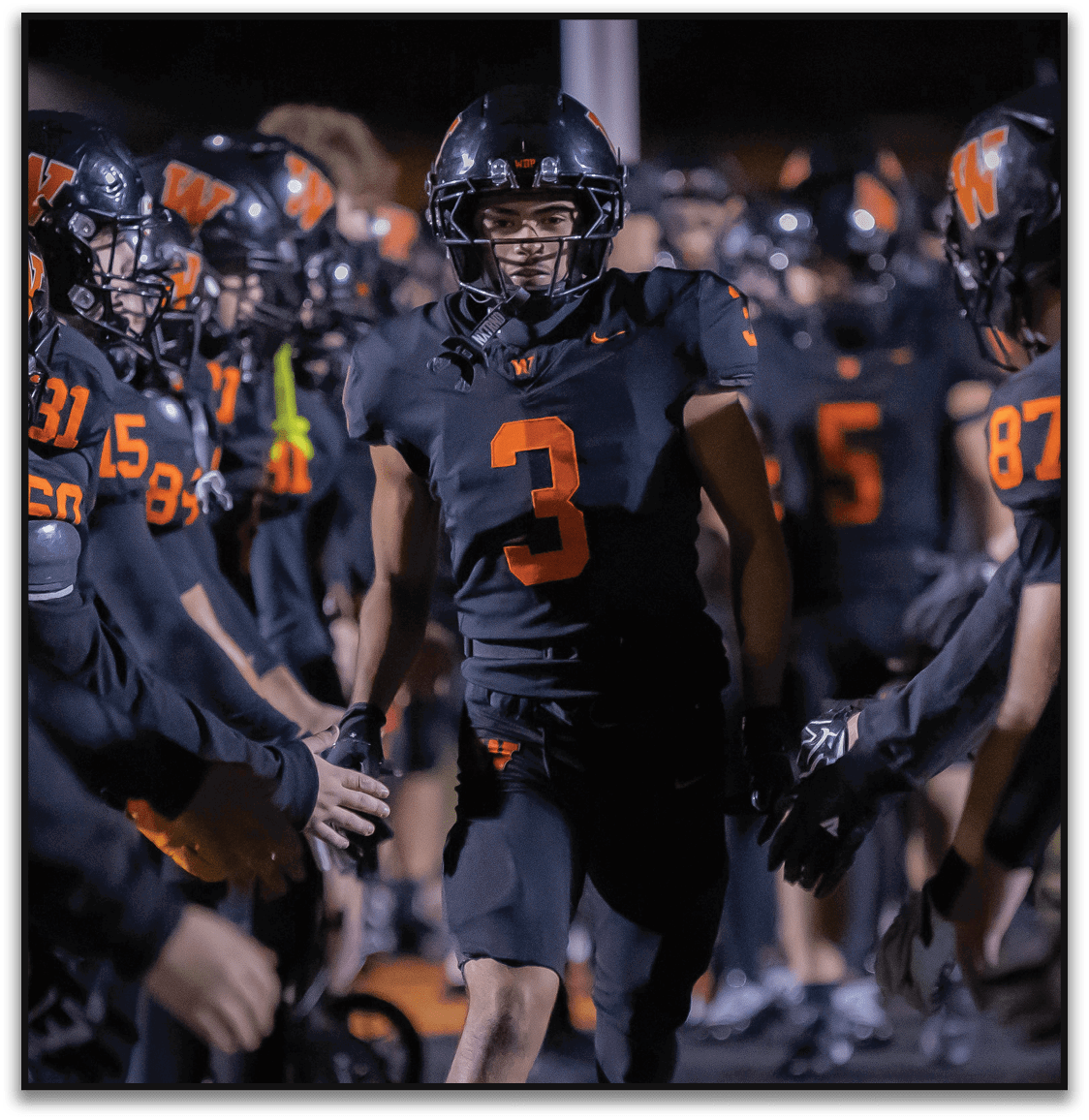 A person wearing a black football uniform with the number 3 is walking through a line of teammates dressed in similar black and orange uniforms giving high fives. Description generated by AI