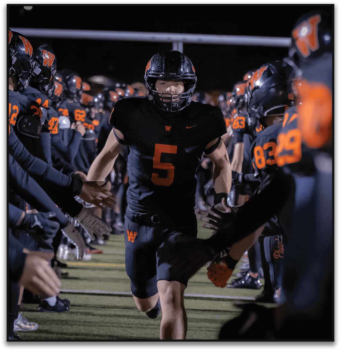 A person wearing a black football uniform with the number five runs through a tunnel formed by teammates giving high fives on a football field at night. Description generated by AI