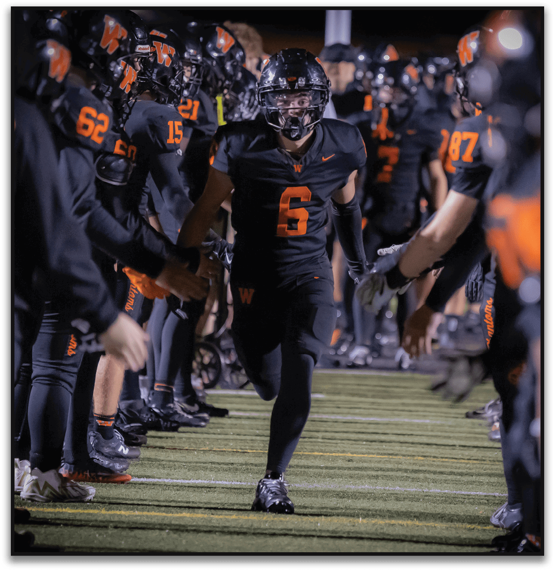 A person wearing a black football uniform with the number six runs through a tunnel formed by teammates in similar black and orange uniforms on a football field at night. Description generated by AI
