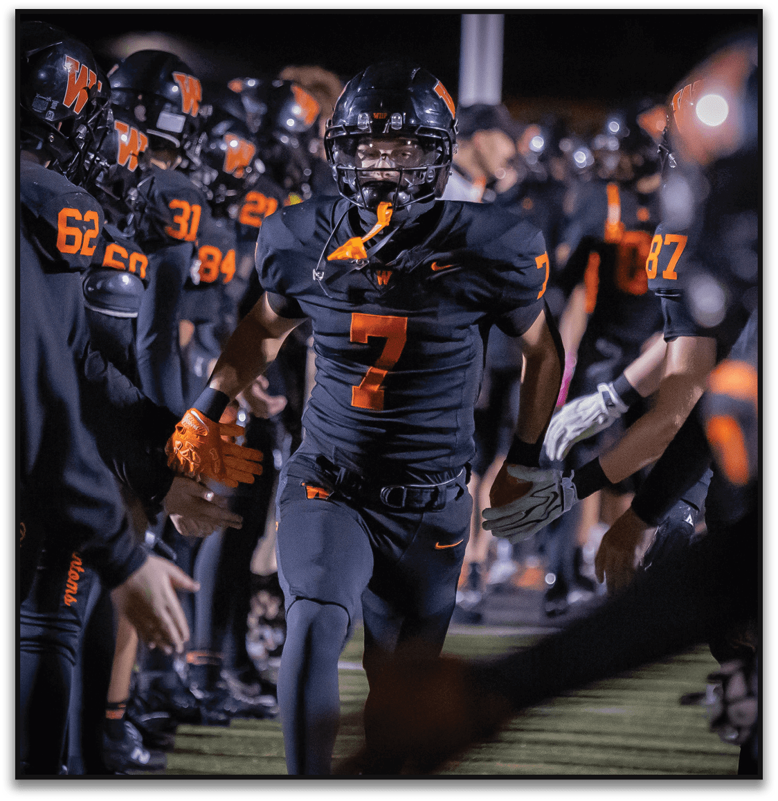 A person wearing a black football uniform with the number seven runs through a line of teammates giving high fives on a field at night. Description generated by AI