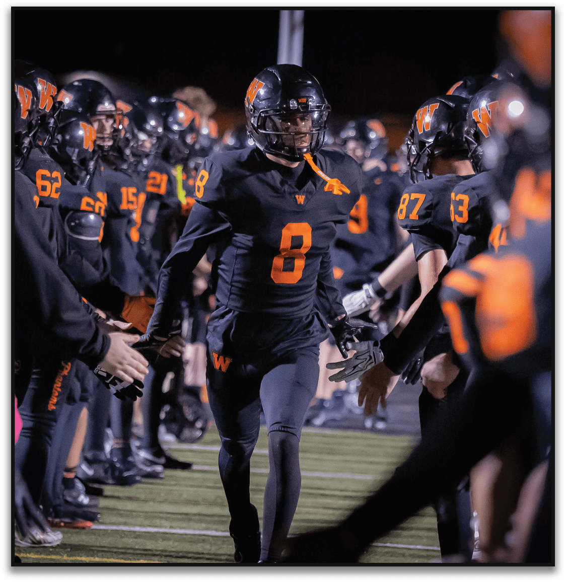 A person wearing a black football uniform with the number 8 is running through a line of teammates who are giving high fives on a football field at night. Description generated by AI