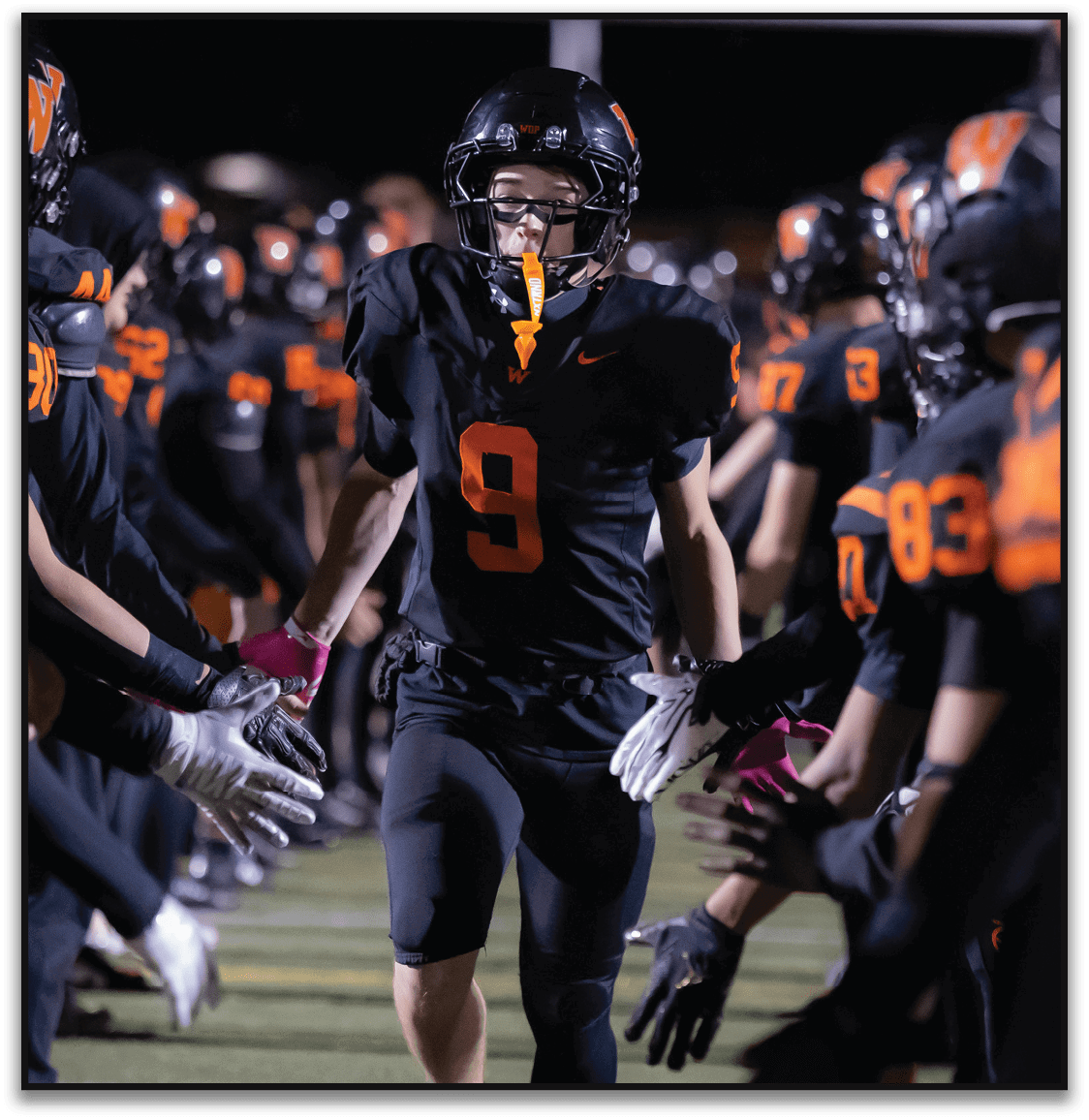 A person wearing a black football uniform with the number nine runs through a tunnel formed by teammates giving high fives on a field at night. Description generated by AI