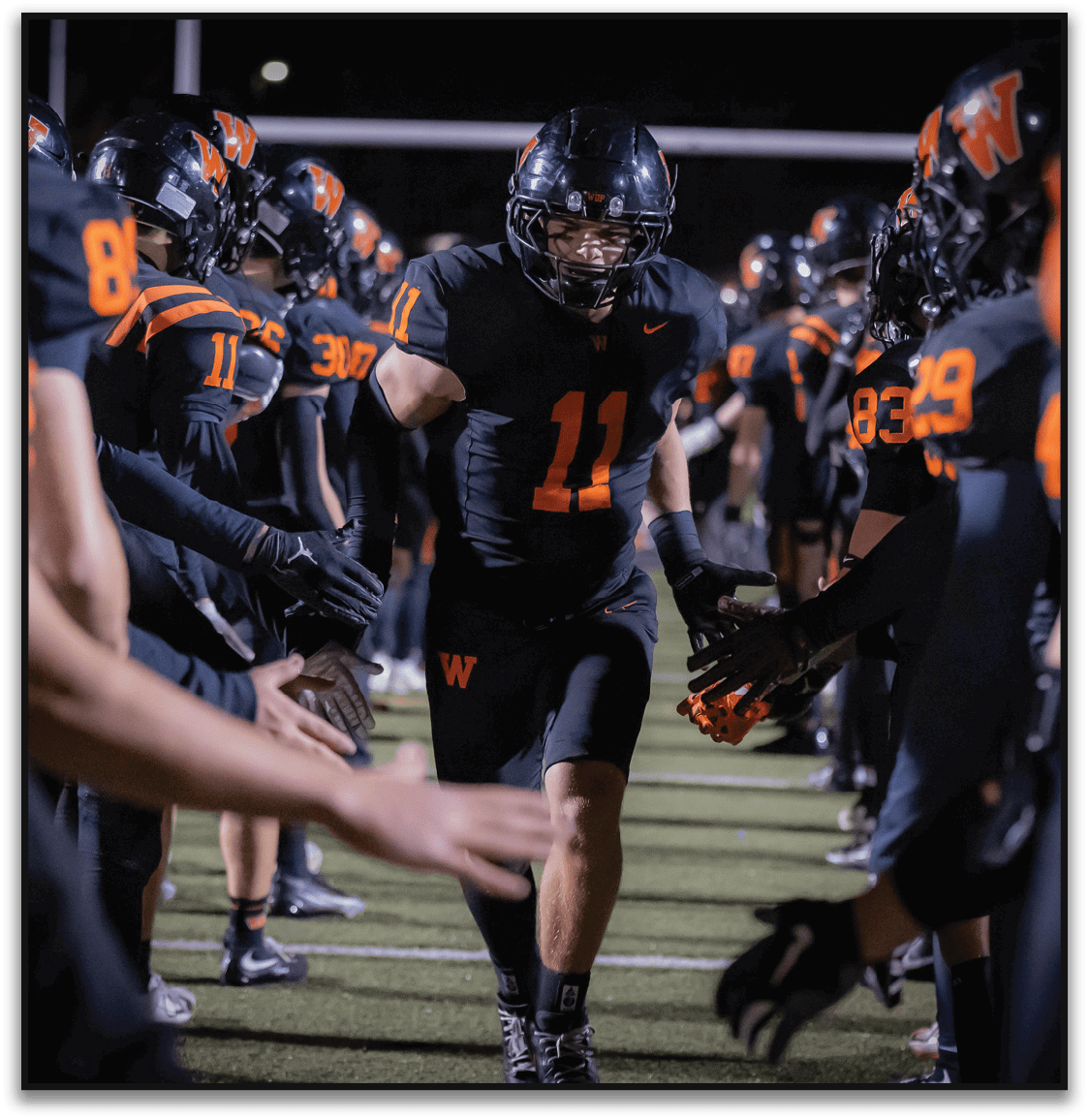 A person wearing a black football uniform with the number 11 is running through a tunnel formed by teammates giving high fives on a lit football field at night. Description generated by AI