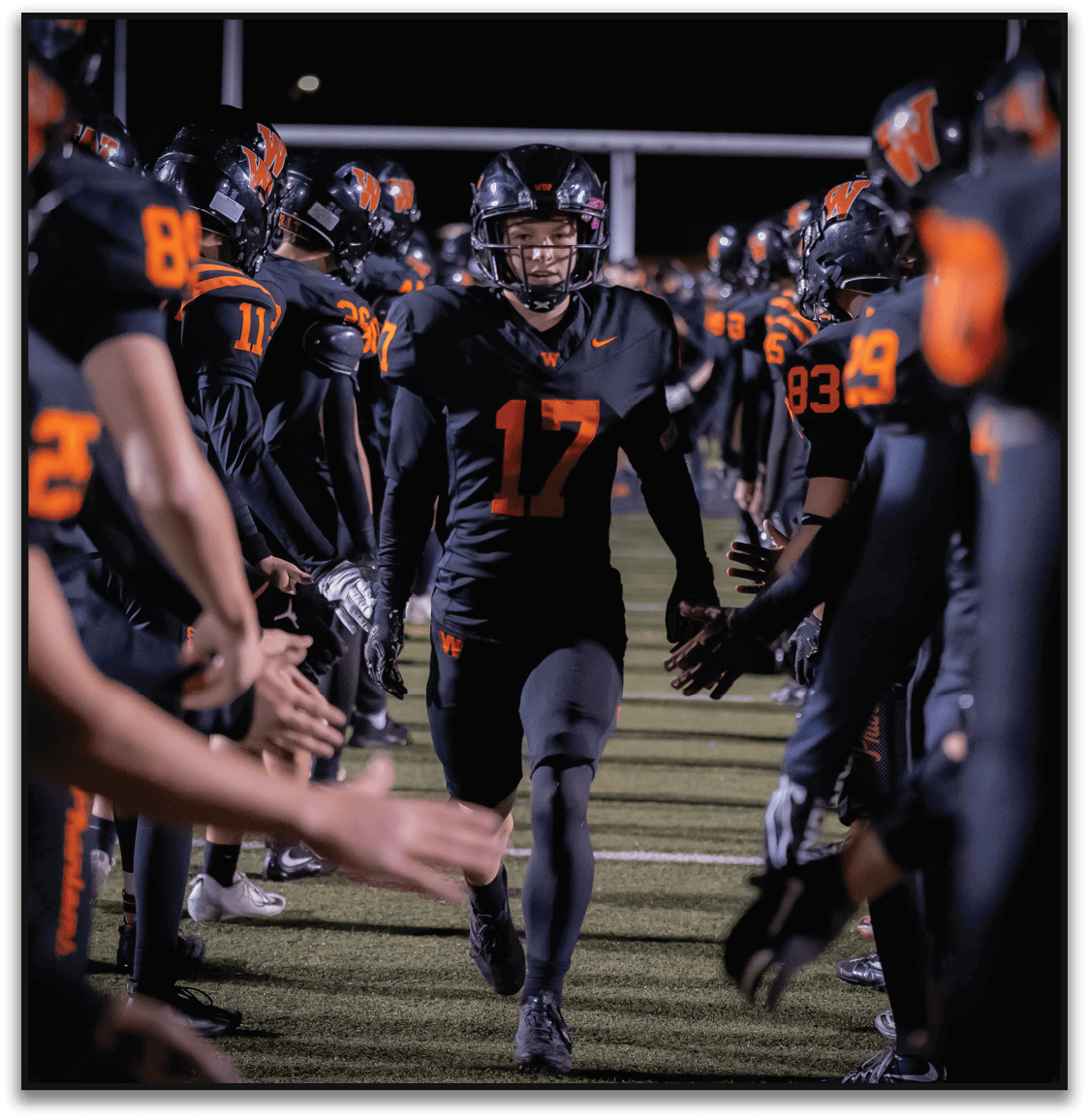 A person wearing a black football uniform with the number 17 is running between two lines of teammates who are giving high fives on a football field at night. Description generated by AI