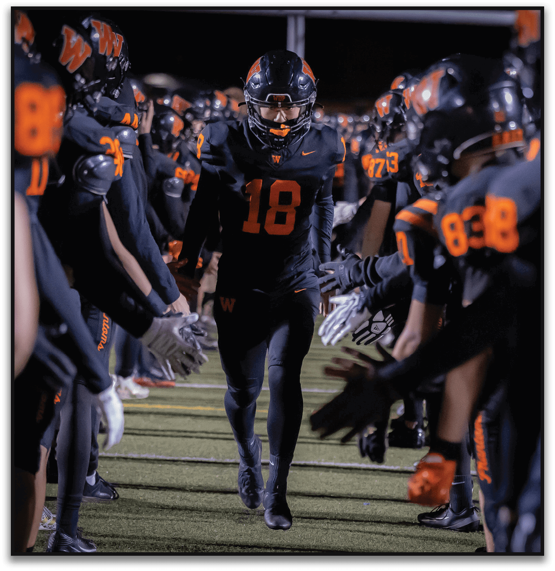 A person wearing a black and orange football uniform with the number 18 walks between two lines of teammates who are reaching out to give high fives on a sports field at night. Description generated by AI