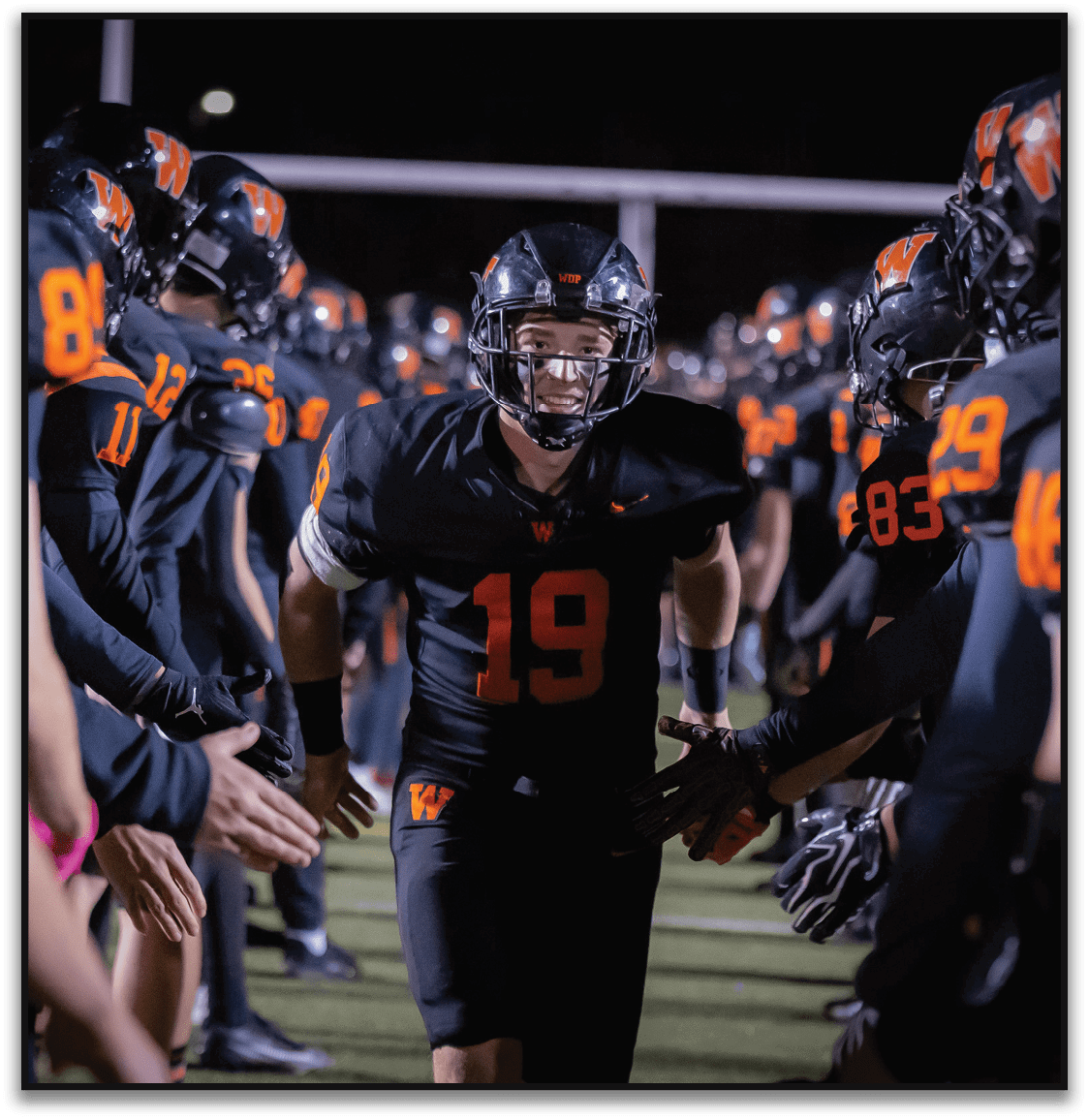 A person wearing a black football uniform with the number 19 is running through a tunnel formed by teammates in similar black uniforms with orange numbers on a football field at night. Description generated by AI