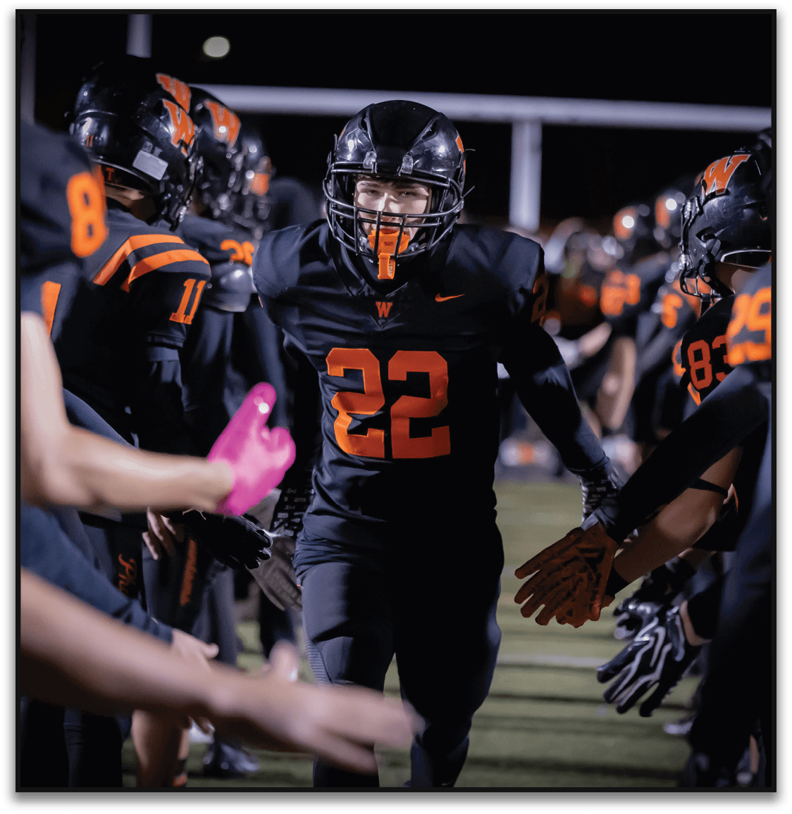A person wearing a black football uniform with the number 22 is running through a line of teammates who are reaching out to give high fives on a lit football field at night. Description generated by AI