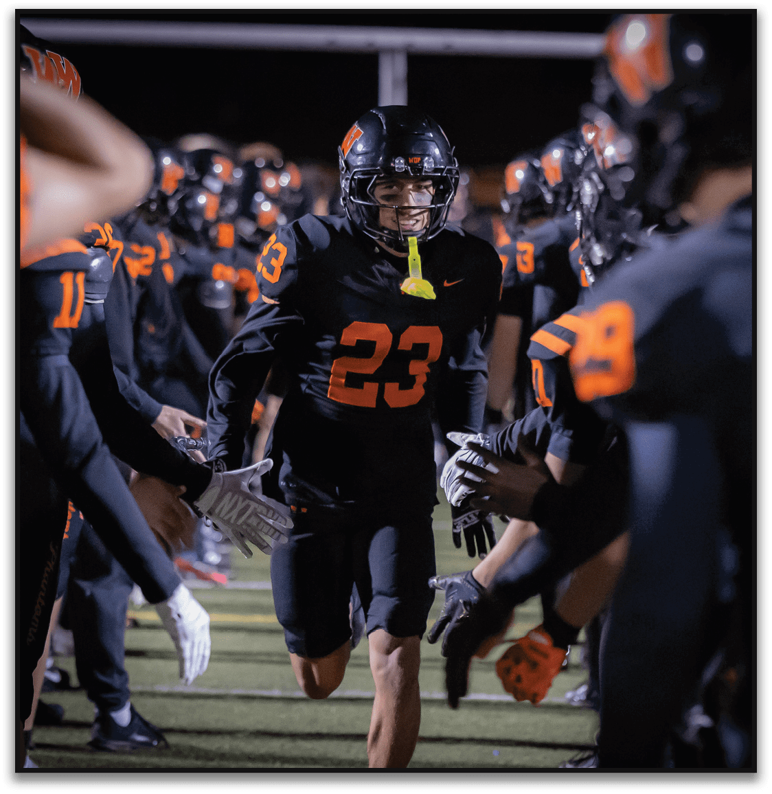 A person wearing a black football uniform with the number 23 runs through a line of teammates who are giving high fives on a football field at night. Description generated by AI