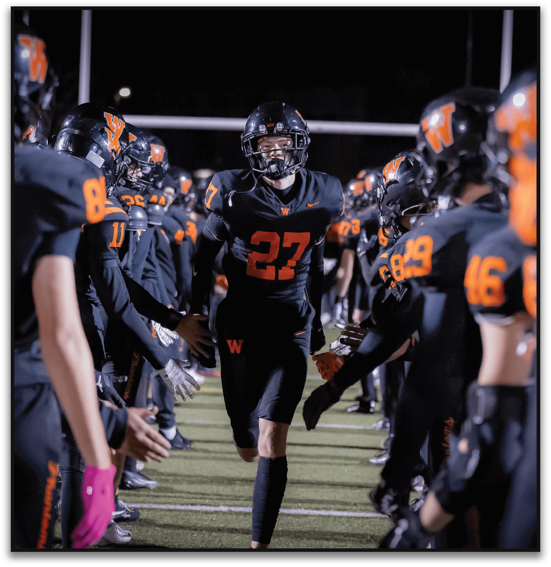 An athlete wearing a black football uniform with the number 27 runs between two lines of teammates in similar black and orange uniforms on a football field at night. Description generated by AI