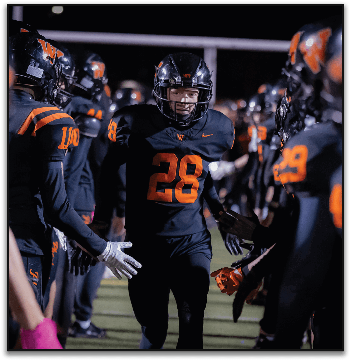 A person wearing a black football uniform with the number 28 is running through a line of teammates who are giving high fives on a field at night. Description generated by AI