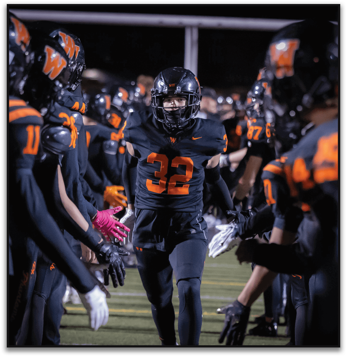 A person wearing a black football uniform with the number 32 is running through a line of teammates dressed in matching black and orange uniforms who are reaching out to give high fives. Description generated by AI