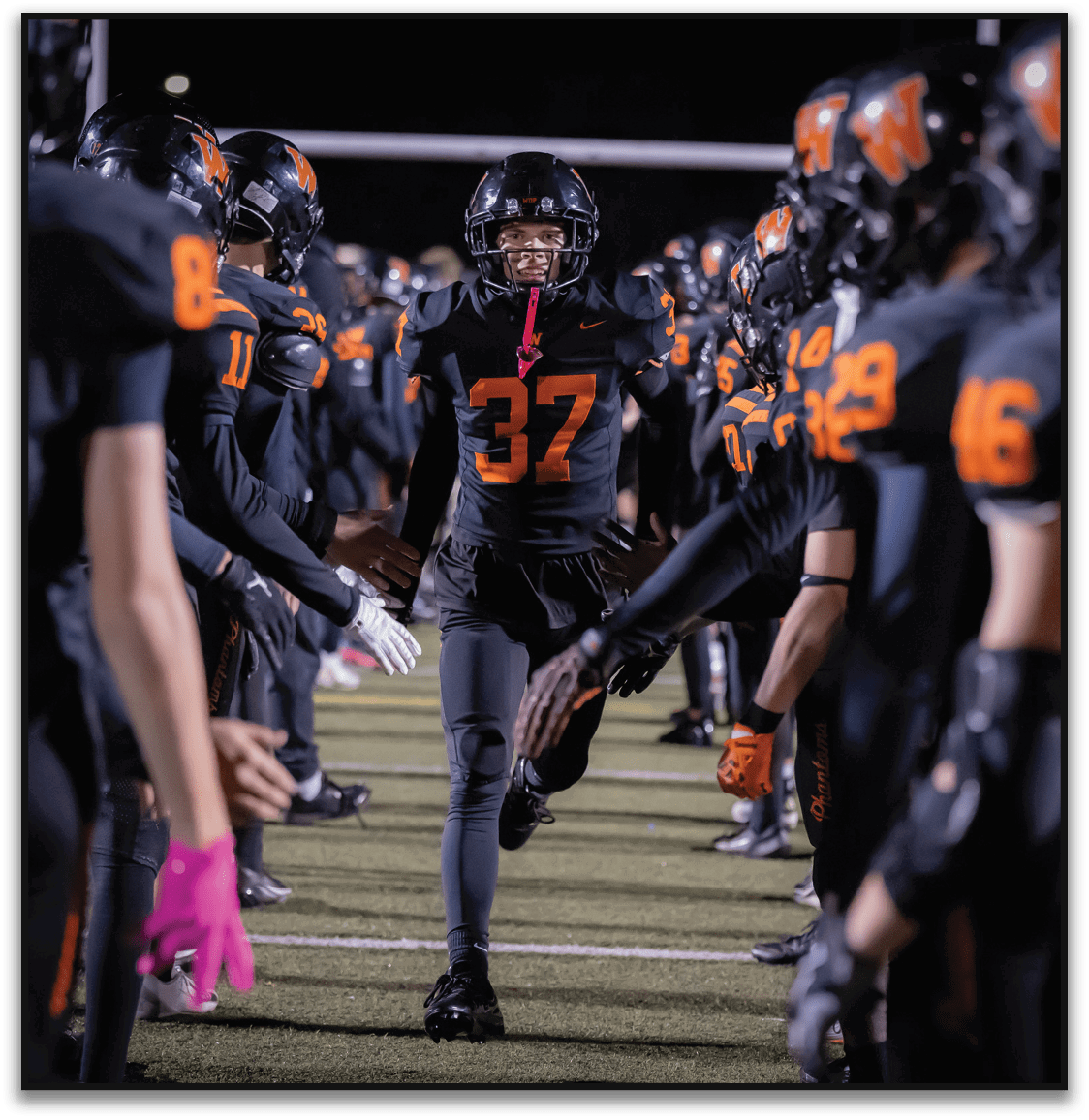 A person wearing a black football uniform with the number 37 runs between two lines of teammates who are clapping and wearing matching black uniforms with orange numbers on a football field at night. Description generated by AI