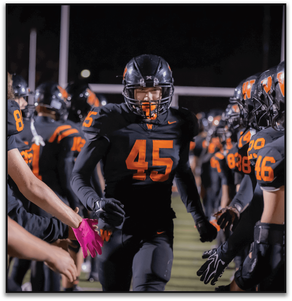 A person wearing a black football uniform with the number 45 is running through a line of teammates who are giving high fives on a football field at night. Description generated by AI