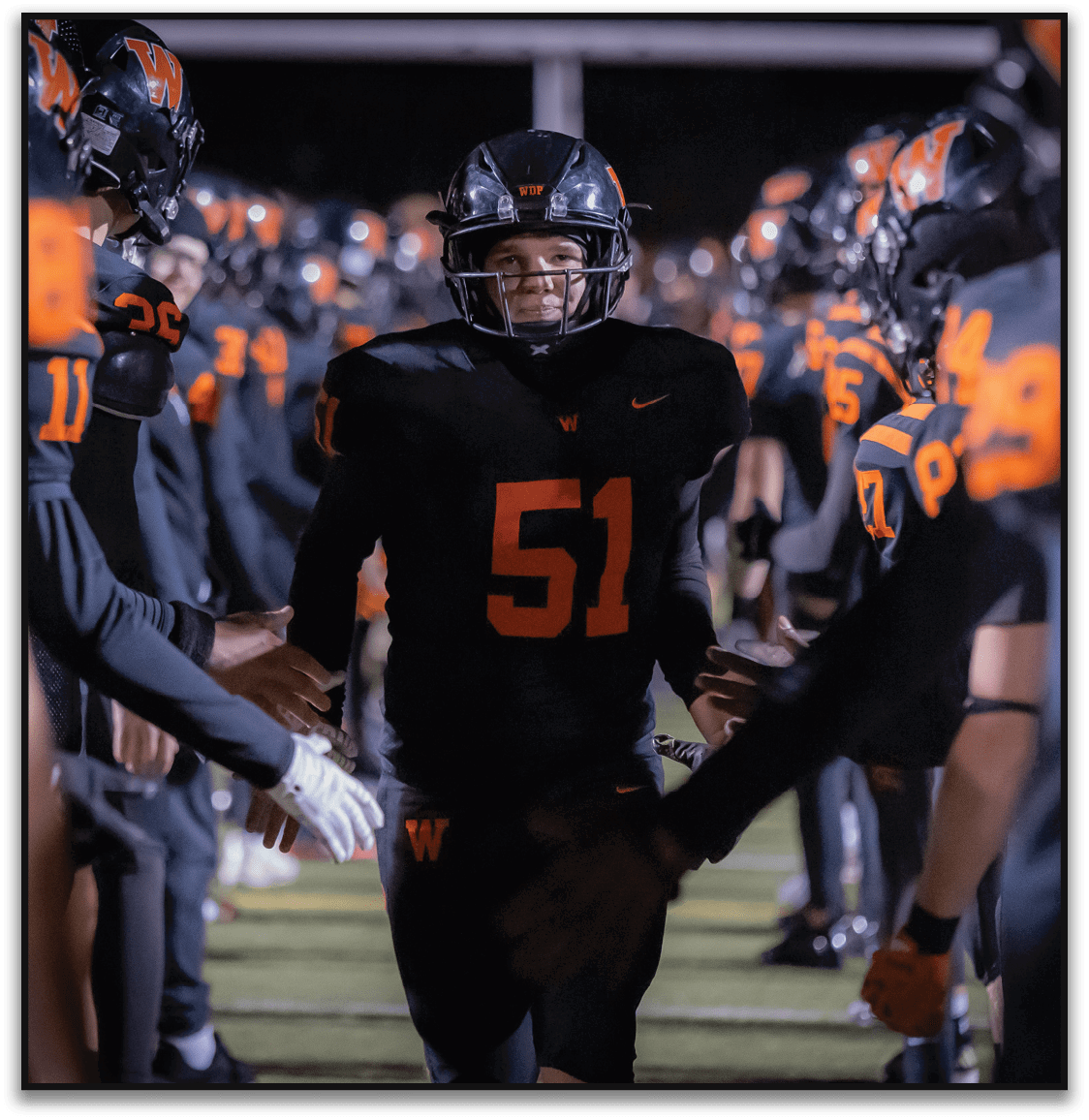 A person wearing a black football uniform with the number 51 is walking through a line of teammates who are reaching out to give high fives on a football field at night. Description generated by AI