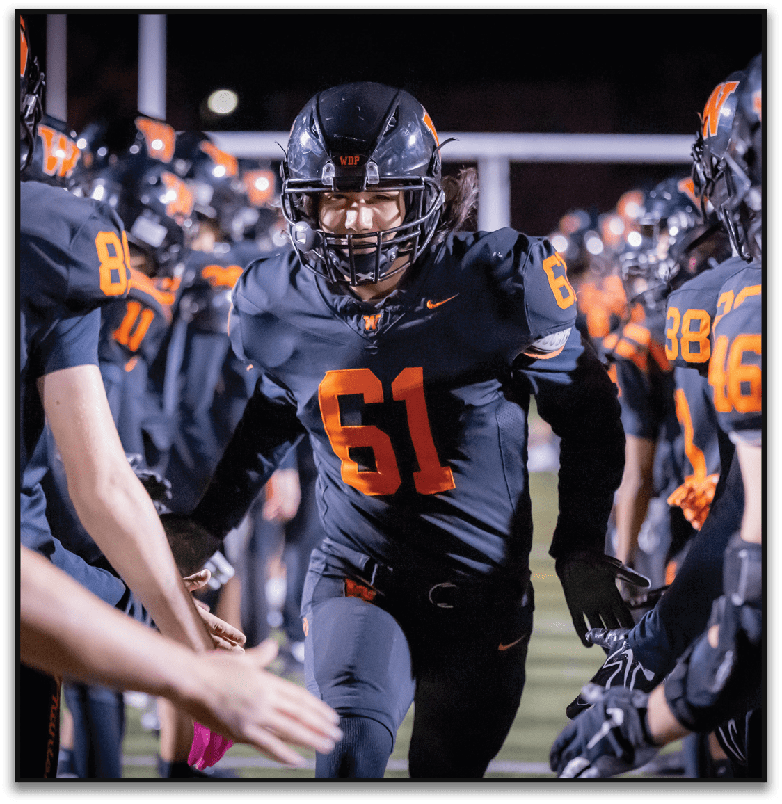 A person wearing a black football uniform with the number 61 is running through a tunnel formed by teammates giving high fives on a lit football field at night. Description generated by AI