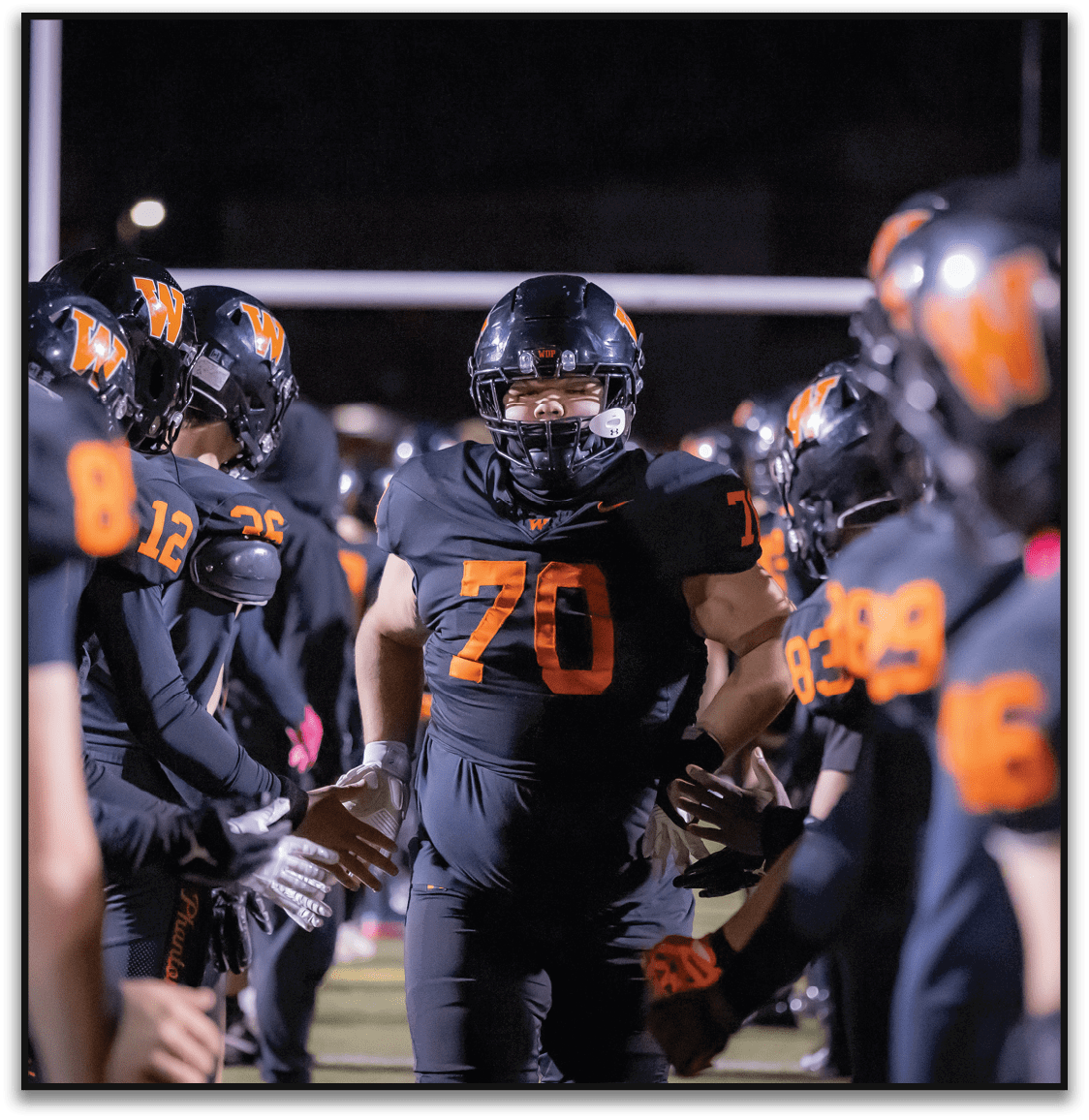 A person wearing a black football uniform with the number 70 is walking through a line of teammates who are giving high fives on a football field at night. Description generated by AI