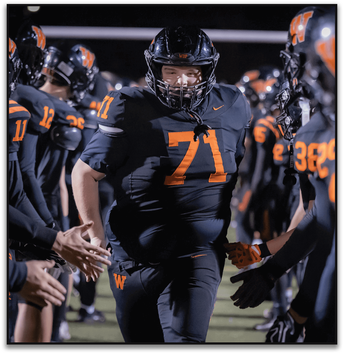 A person wearing a black football uniform with the number 71 is running through a line of teammates who are giving high fives on a football field at night. Description generated by AI