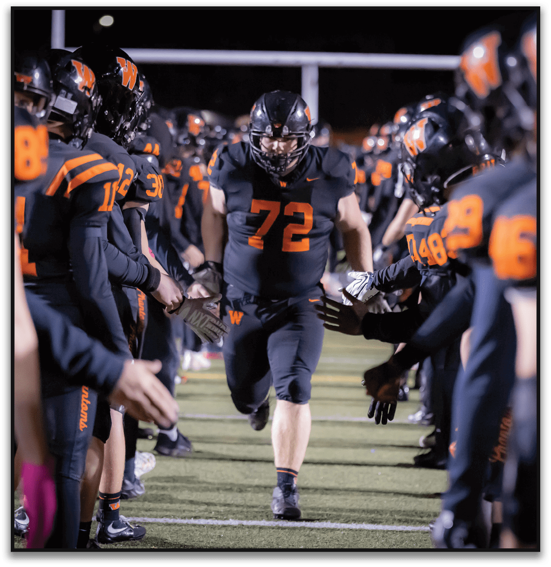 A person wearing a black football uniform with the number 72 is running down a line of teammates in similar black and orange uniforms who are reaching out to give high fives on a football field at night. Description generated by AI